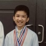 A young boy smiling, wearing a light blue shirt and tie, with several medals around his neck, standing in front of a dark door.
