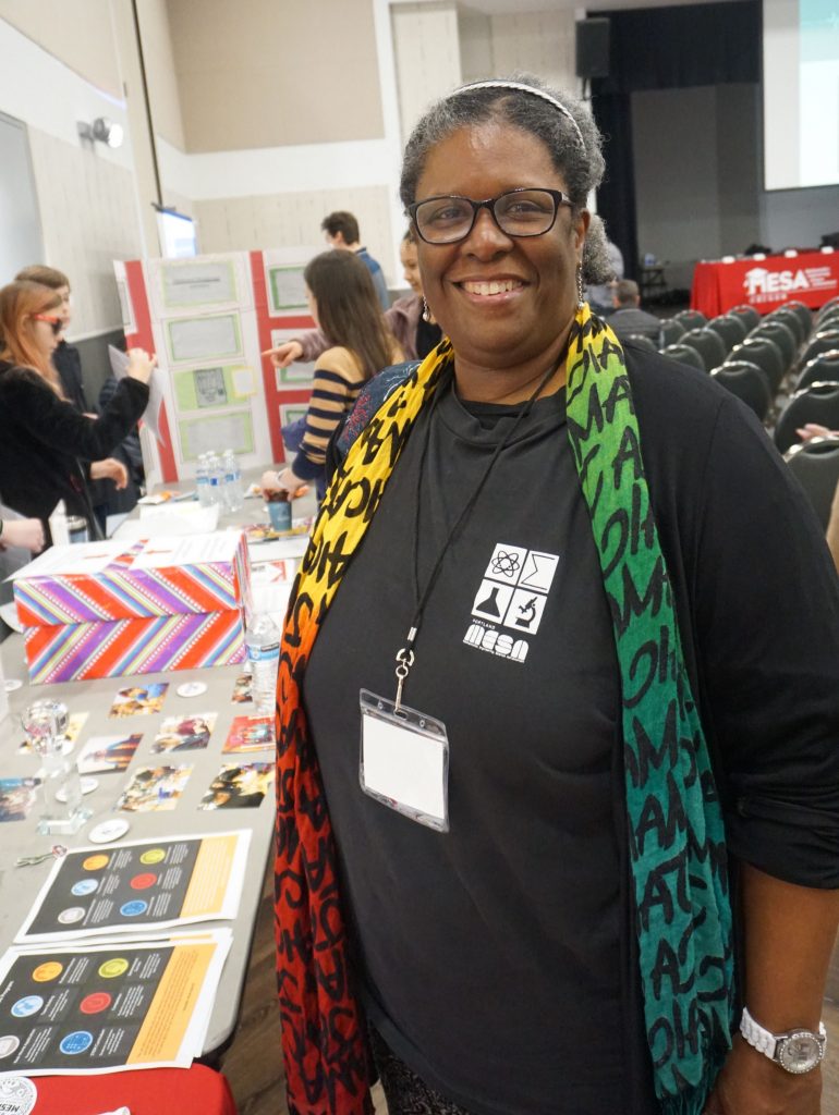 A smiling woman with gray hair and glasses stands indoors near a display table. She wears a black shirt with a logo, a colorful scarf, and a name badge. People and informational posters are visible in the background.
