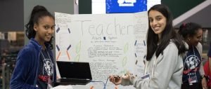 Two girls stand in front of a science fair project display board titled Teacher Alert System, smiling at the camera. One holds a laptop, and the other holds a paper. Another person is visible in the background.