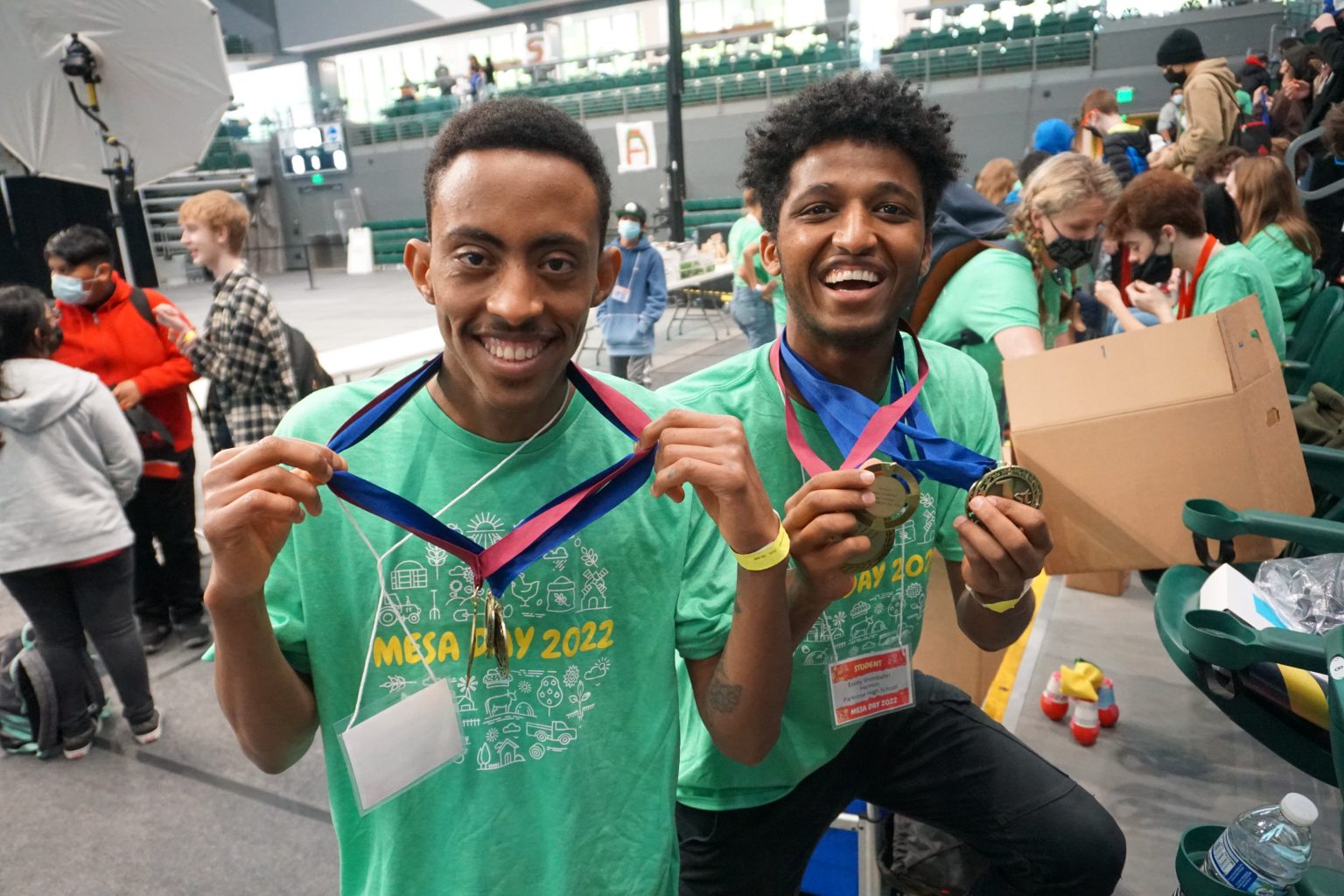 Two smiling young men wearing green MESA Day 2022 shirts proudly show off medals around their necks in a busy indoor event space with other participants and spectators in the background.