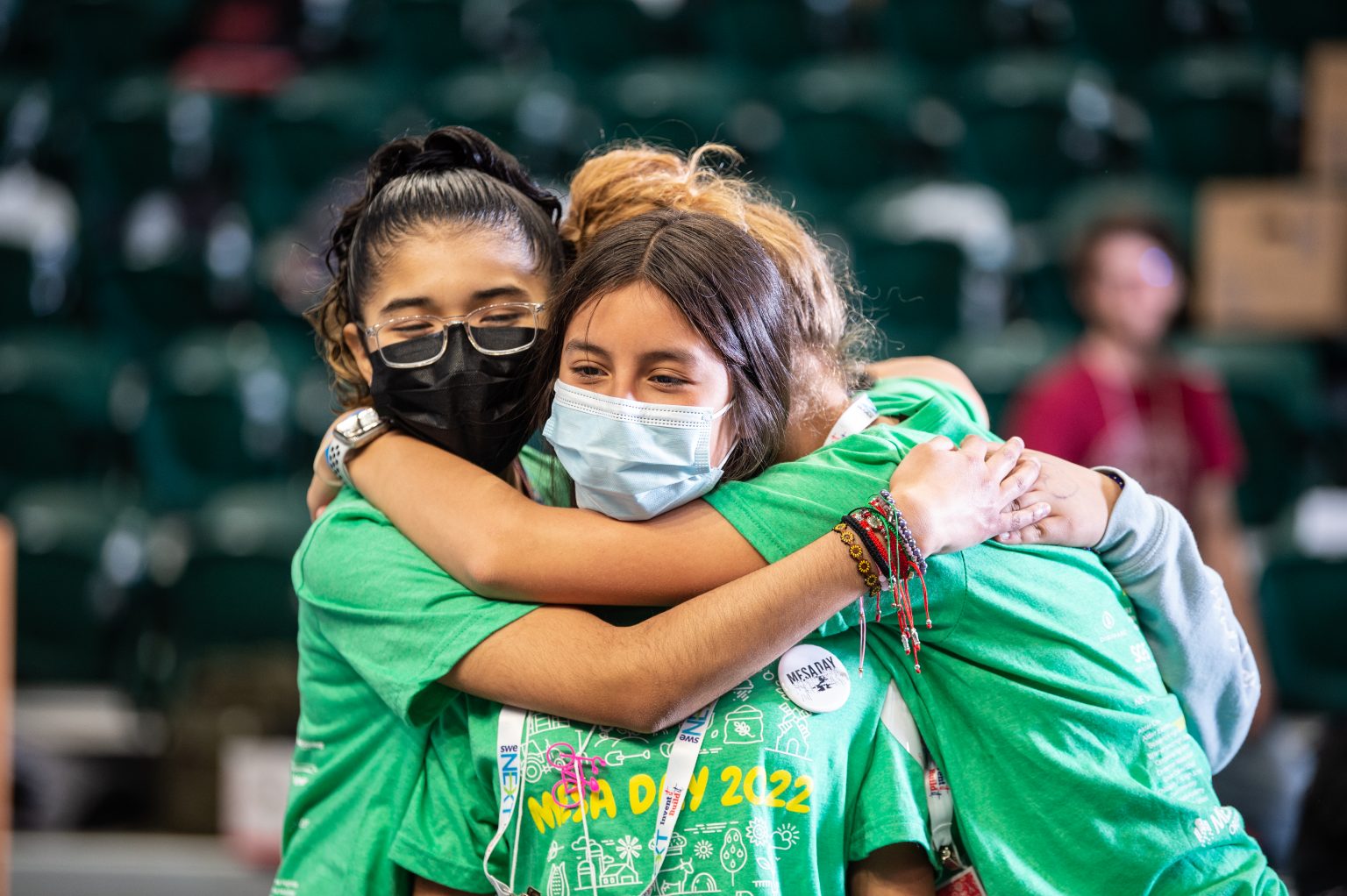 Four young people wearing green MECA Day 2022 shirts and face masks embrace in a group hug, showing friendship and support. The background is blurred, focusing on their joyful expressions.