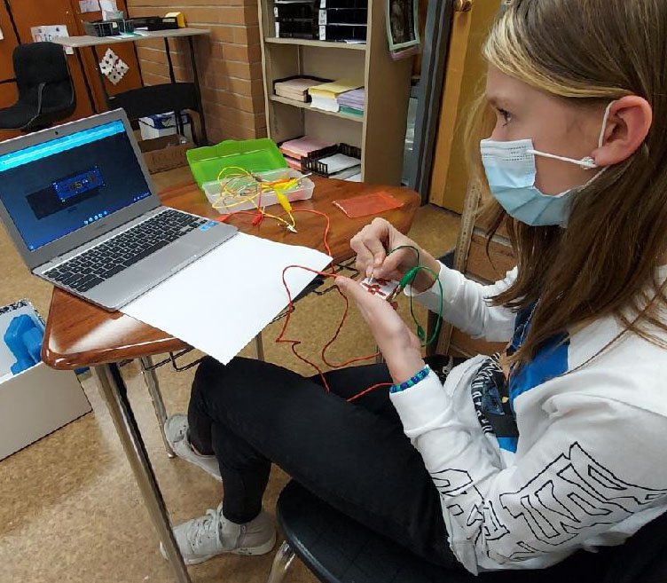 A student wearing a mask sits at a desk, holding wires connected to a small circuit board while working on a project with a laptop in a classroom.