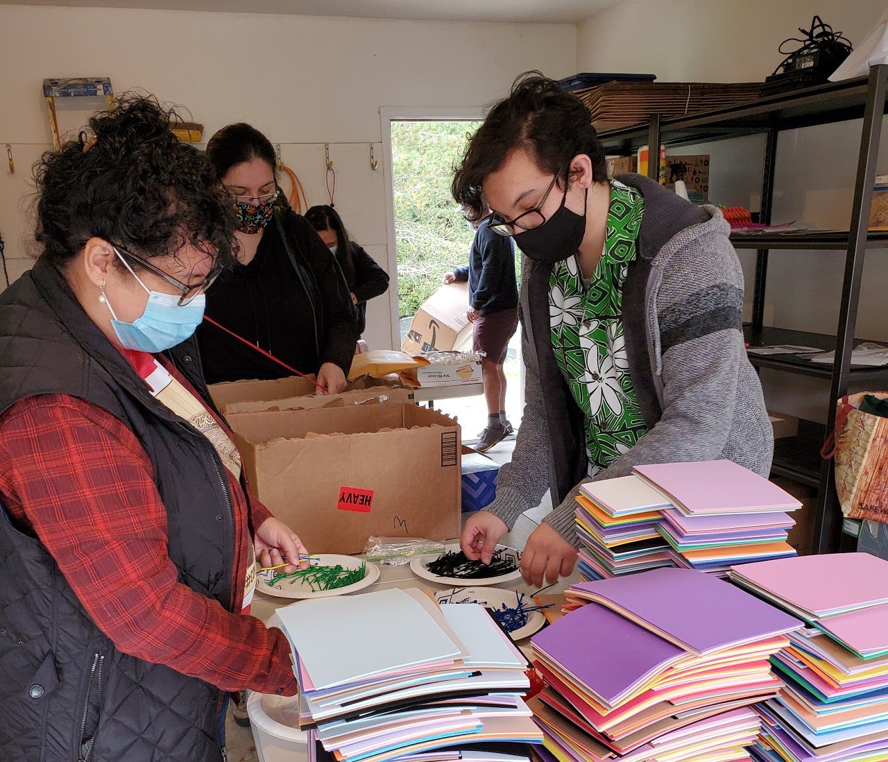 Four people wearing masks are assembling and sorting stacks of colorful paper and supplies on tables in a room. Shelves and paper bags are visible in the background.