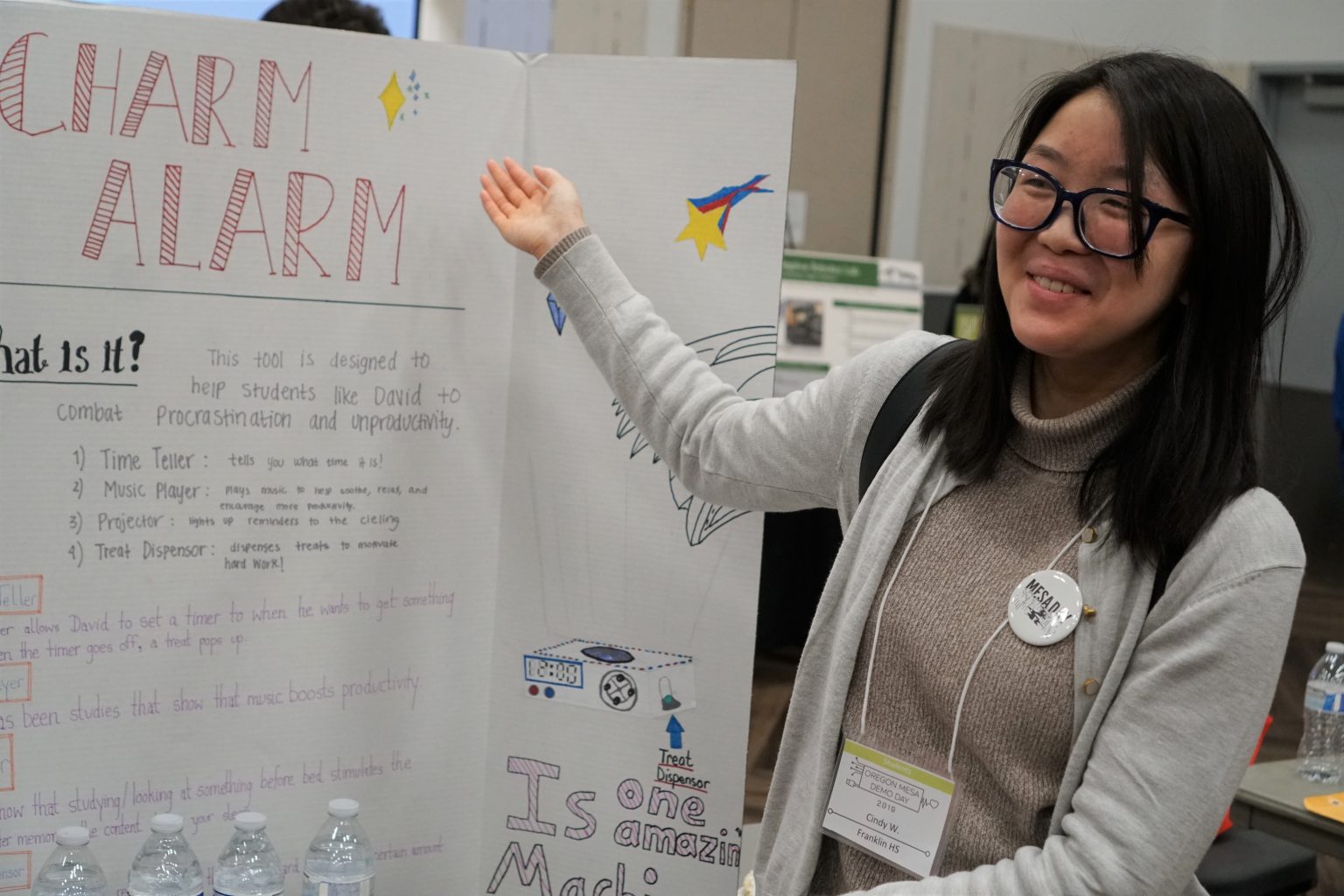A young woman smiles and gestures toward a science fair display titled CHARM ALARM. She wears glasses, a gray turtleneck, and a lanyard with badges. The poster explains a tool to help with procrastination and productivity.