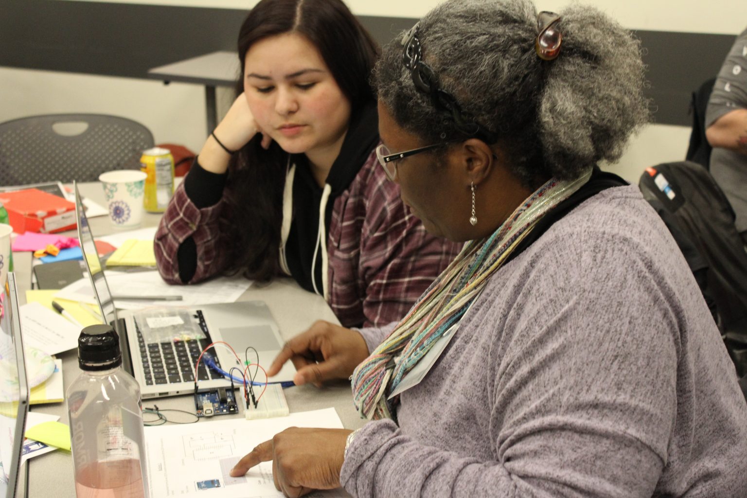 Two women sit at a table working on a project with a laptop, electronic components, and papers. One woman points at a document while the other listens attentively.
