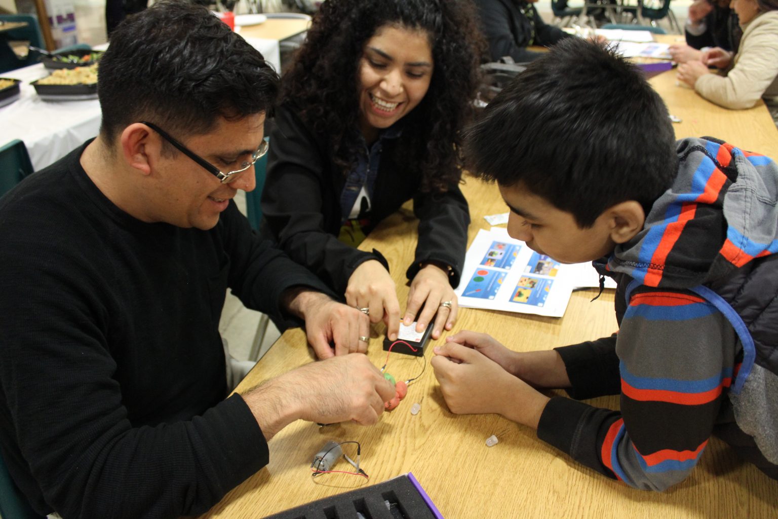 Three people—an adult man, a woman, and a boy—sit at a table, smiling and working together on an electronics project with wires and small components. Instruction sheets are visible on the table.