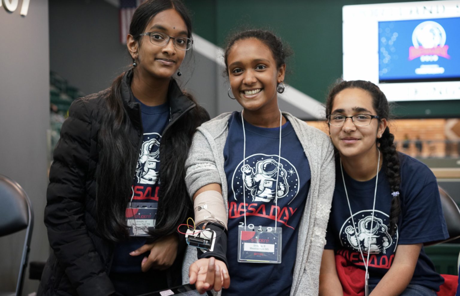 Three girls wearing matching NASA t-shirts smile at the camera; one of them shows a homemade electronic device attached to her arm. They appear to be at a science or robotics event.