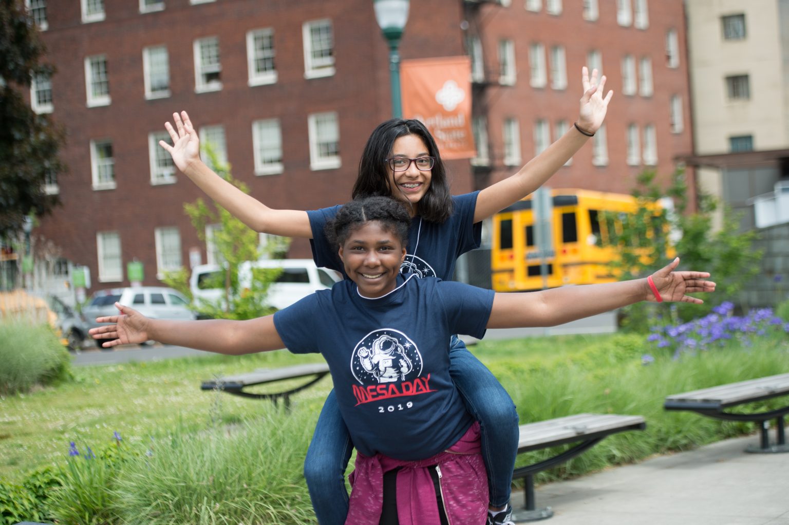 Two smiling kids pose playfully outdoors. One is giving the other a piggyback ride, both have arms outstretched like airplane wings. They are in front of a brick building with benches, greenery, and a yellow school bus in the background.