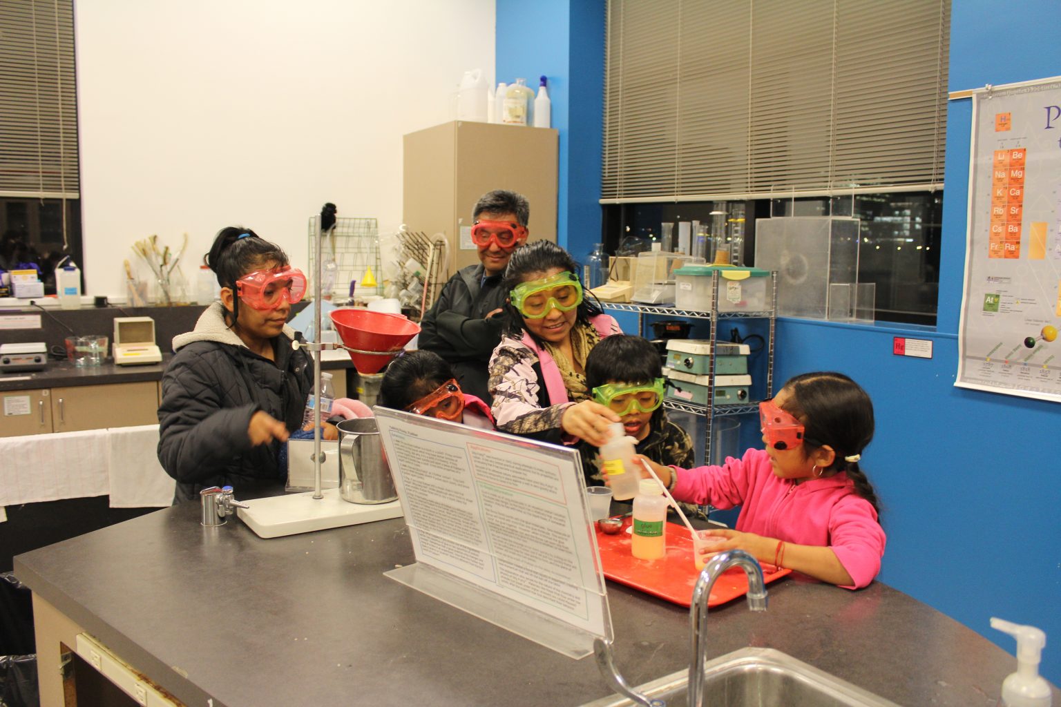 A group of children and two adults wearing colorful safety goggles conduct a science experiment together at a lab table, surrounded by equipment and a periodic table poster on the wall.