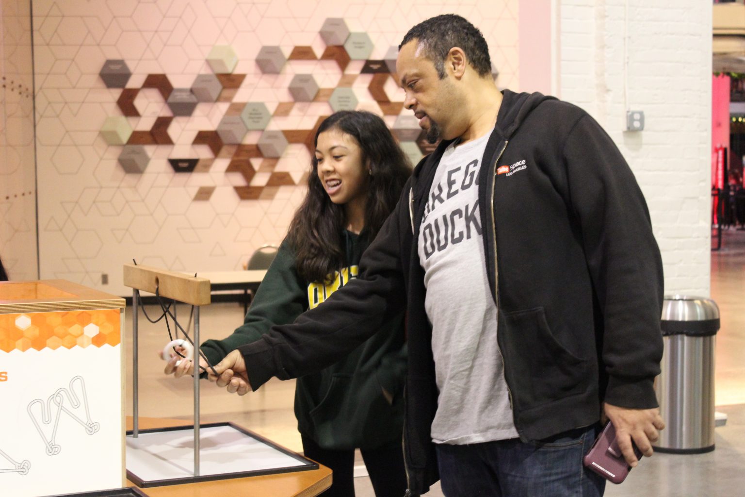 A man and a girl are interacting with a hands-on science exhibit indoors. The man holds a rod while the girl smiles and looks on. The background features a geometric wall design and a trash can.