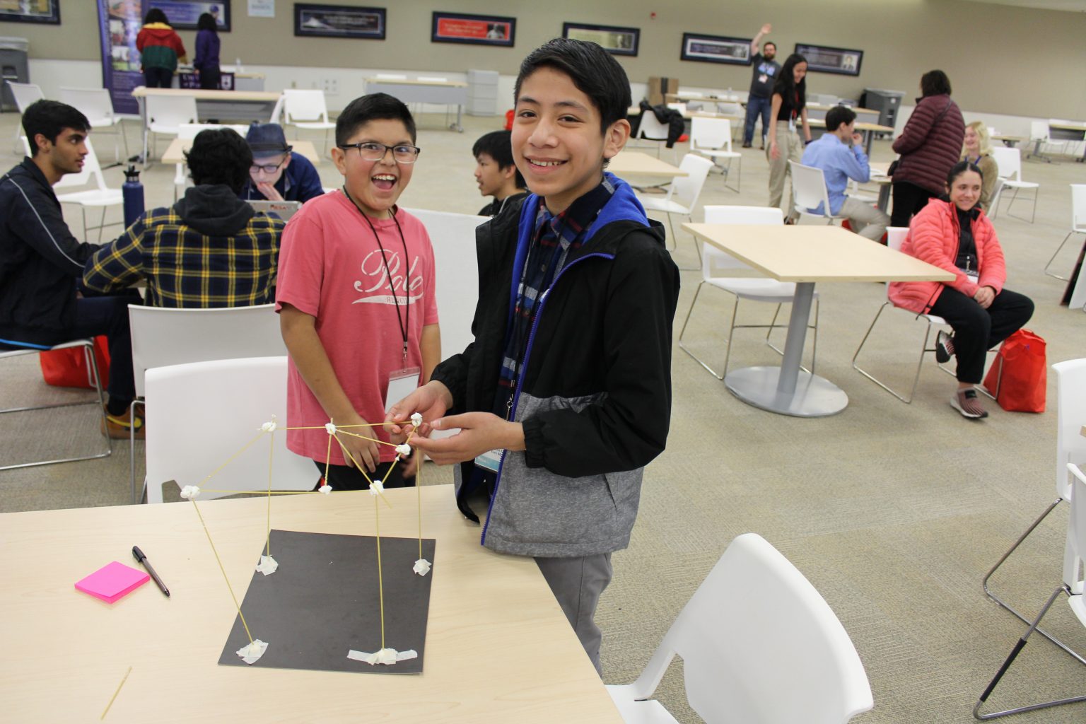 Two boys smile while building a structure out of spaghetti and marshmallows at a table in a large, well-lit room with other people and groups working in the background.
