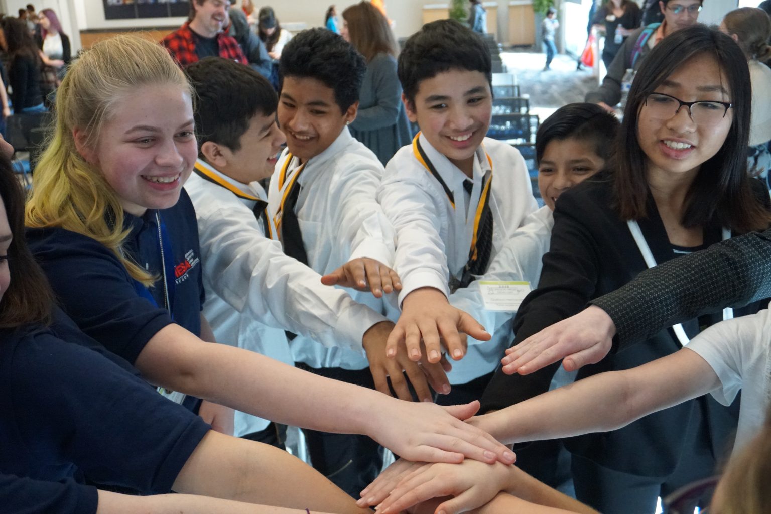 A group of smiling students standing in a circle, each reaching one hand into the center to stack their hands together in a gesture of teamwork and unity.