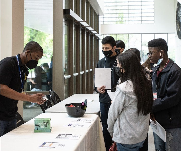 A group of young people wearing masks stands at a registration table indoors, where a staff member is handing something to one of them. Papers and a bowl with items are on the table. Large windows are in the background.