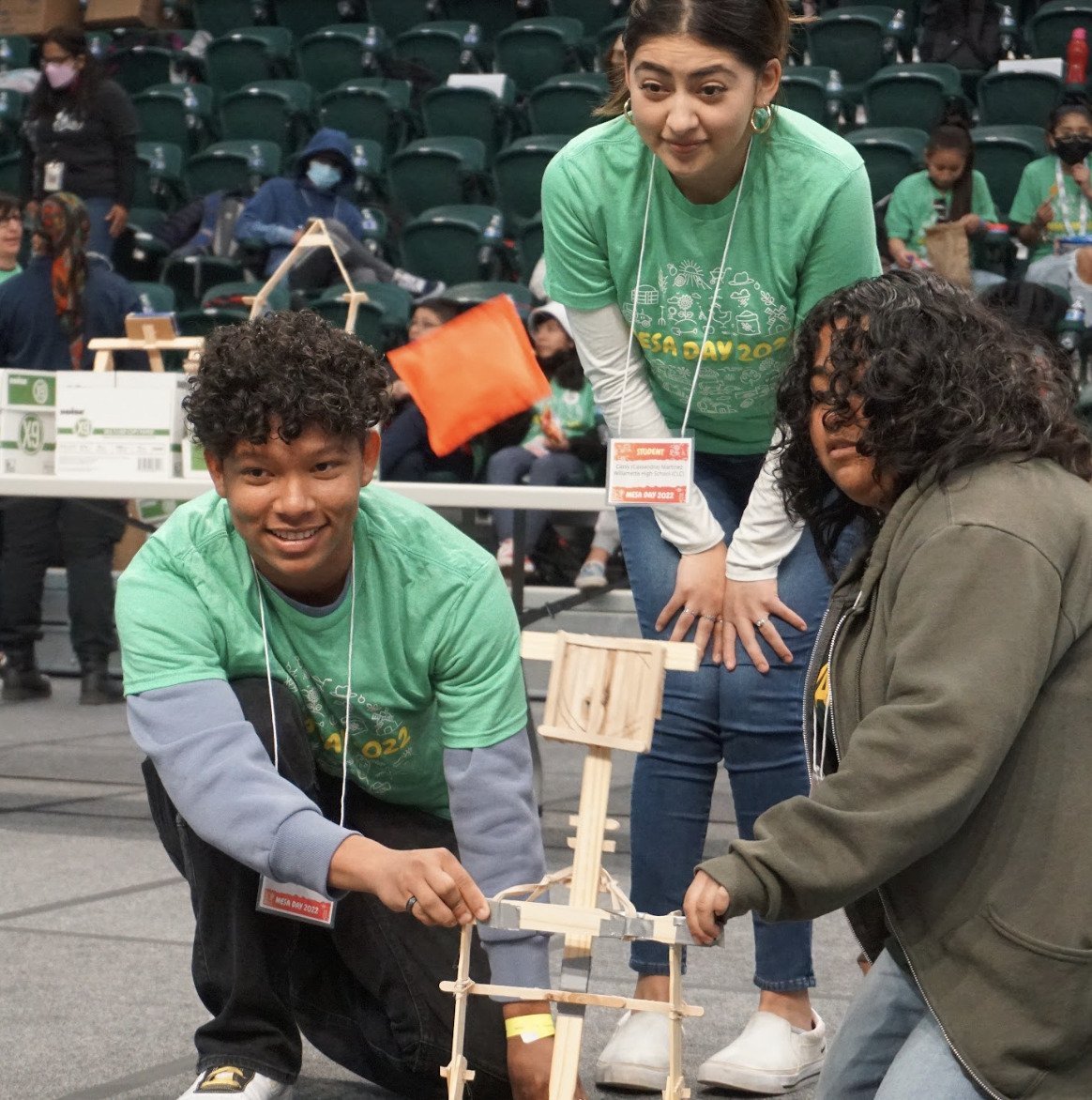 Three students in green shirts kneel and crouch on a gym floor, working together to build a small wooden structure. Other people and rows of green chairs are visible in the background.