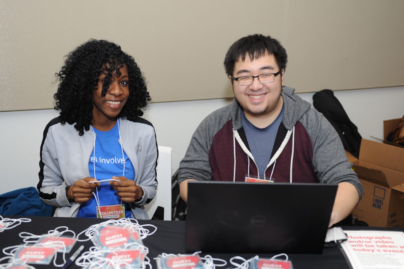 Two volunteers sit at a table with name badges and lanyards. One woman, in a blue shirt, smiles while holding lanyards. A man next to her, wearing glasses, smiles at a laptop. A cardboard box is in the background.