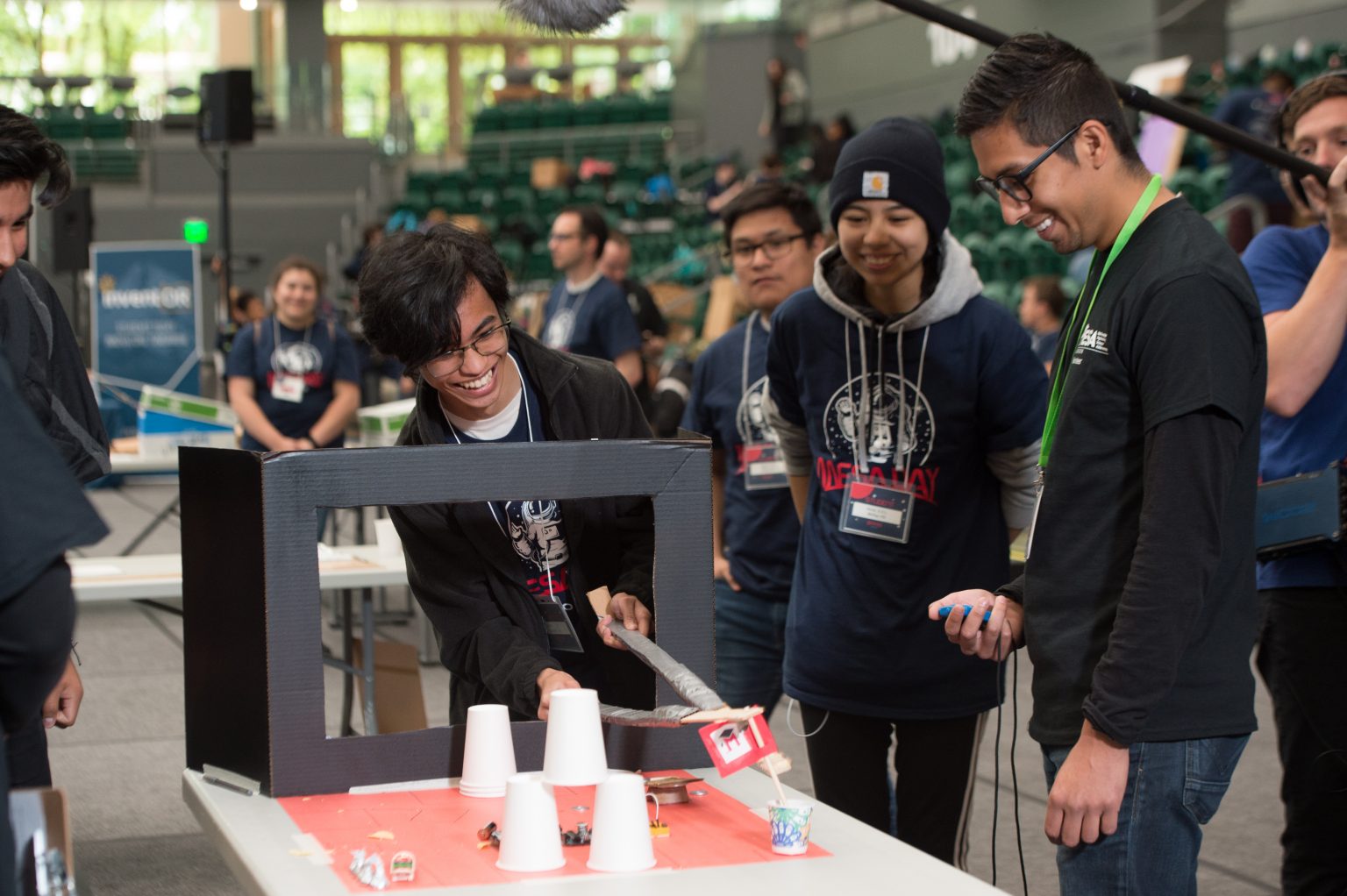 Several young adults gather around a table, smiling and engaged in a hands-on activity involving a homemade contraption, cardboard, and cups; others watch and film in a large indoor venue.