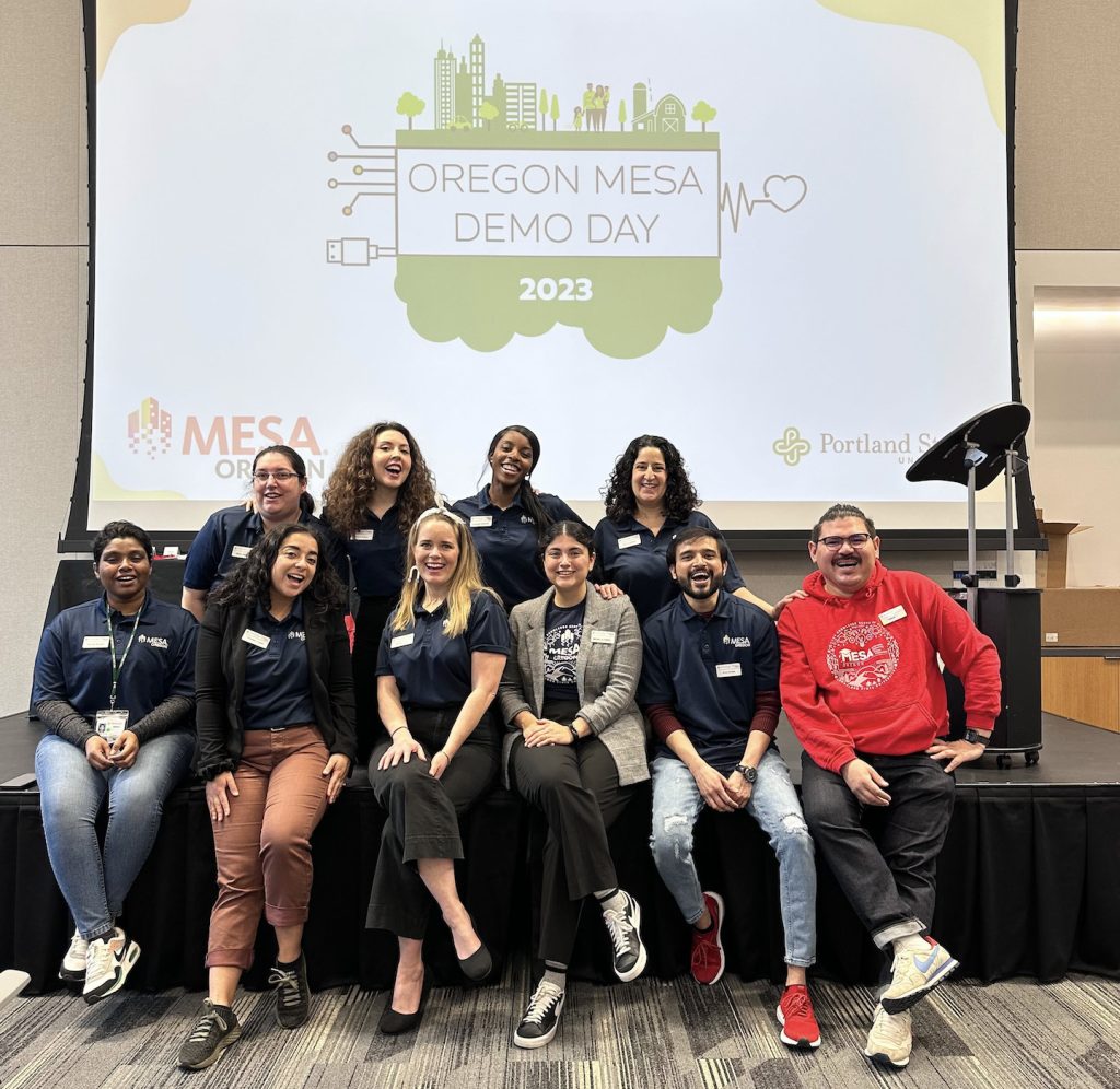 A group of ten people, some seated and some standing, smile for a photo in front of a screen displaying Oregon MESA Demo Day 2023 at an indoor event, captured as part of the demo day 2023 photo gallery.