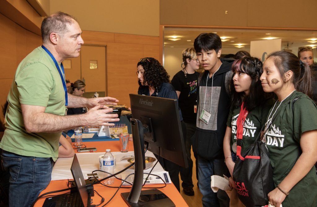 A man speaks animatedly to a group of four students at a table with a computer monitor and informational materials in a bright indoor setting during the MESA Day 2023 photo gallery. The students listen attentively.