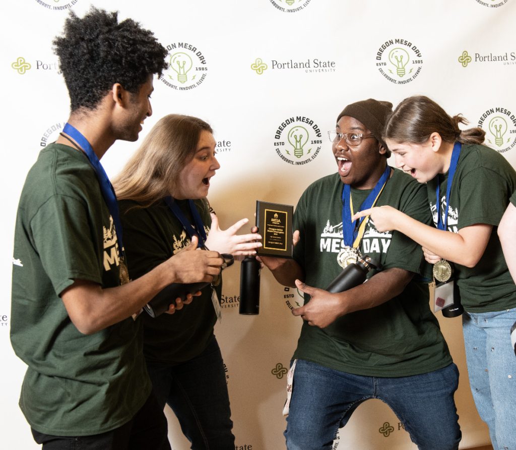 Four excited young people in matching green shirts and medals celebrate while holding a plaque and pointing at it, standing in front of a Portland State University and MESA Day 2023 photo gallery banner.