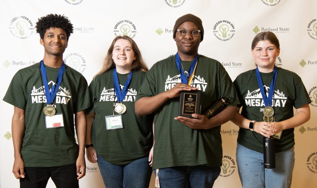 Four students stand side by side, smiling and wearing green MESADay 2024 t-shirts and medals. One holds a plaque, another holds a black water bottle. A branded backdrop is behind them—see more in the MESA Day 2023 photo gallery.