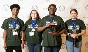 Four students stand side by side, smiling and wearing green MESADay 2024 t-shirts and medals. One holds a plaque, another holds a black water bottle. A branded backdrop is behind them—see more in the MESA Day 2023 photo gallery.