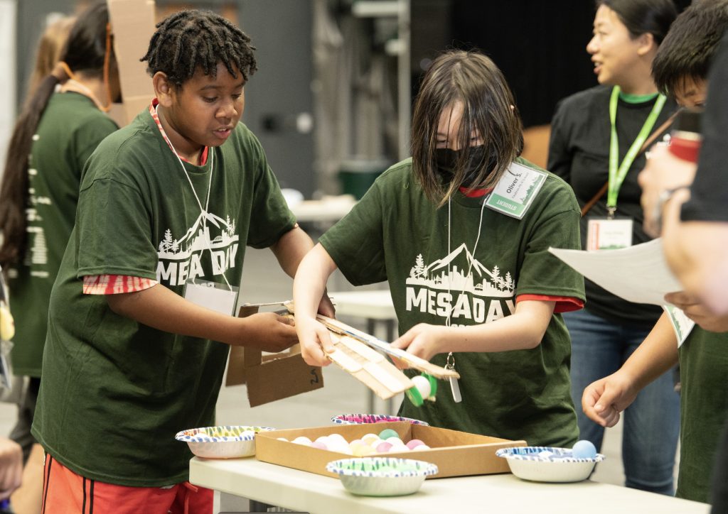 Children wearing green Mesa Day shirts work together on a science project at a table, handling a cardboard structure and colorful plastic eggs, while others look on and hold papers. See more moments in the MESA Day 2023 photo gallery.