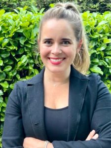 A woman with light skin and long blonde hair in a ponytail stands in front of green bushes, smiling. She is wearing a black blazer, hoop earrings, and a necklace, with her arms crossed.