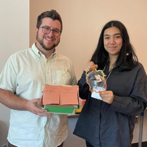 Two people stand indoors, smiling at the camera. Launching into the New School Year, the man on the left holds a cardboard model with handwritten notes, while the woman on the right holds a craft project with orange trim and a clear center.