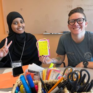 Two people sit at a table covered with craft supplies, smiling at the camera as they prepare for launching into the new school year. One holds up a colorful card, while the other flashes a peace sign. A whiteboard and baskets of supplies are in the background.