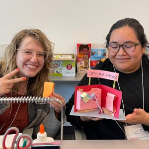 Two smiling people sit at a table with craft supplies, launching into the new school year. One holds a yellow note, and the other shows off a pink “communal mailbox” model. Snack boxes in the background add to their happy, engaged craft session.