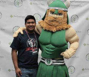 A smiling student, wearing a black T-shirt and red name tag, stands next to a green Viking mascot at an event with a Portland State University backdrop, celebrating the impact of The Oregon MESA Alumni Endowed Scholarship.