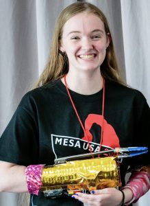 A smiling young woman with long light brown hair wears a black Mesa USA T-shirt and a red lanyard. Representing MESA Alumna Jamie's Story, she holds a gold-colored robotic arm with pink and blue accents. A gray curtain is in the background.