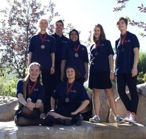Seven people wearing matching navy shirts and medals, including MESA Alumna Jamie, pose outdoors on large rocks, smiling at the camera. Trees and greenery are visible in the bright background, celebrating Jamie's story of success.