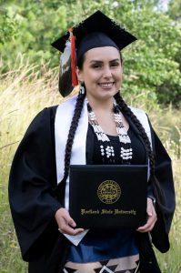 A woman wearing a graduation cap and gown with traditional beadwork stands outdoors, smiling and holding a diploma folder from Portland State University. Green foliage and tall grass are in the background, highlighting Changing Lives Through Engineering: Brook Thompson's Story.