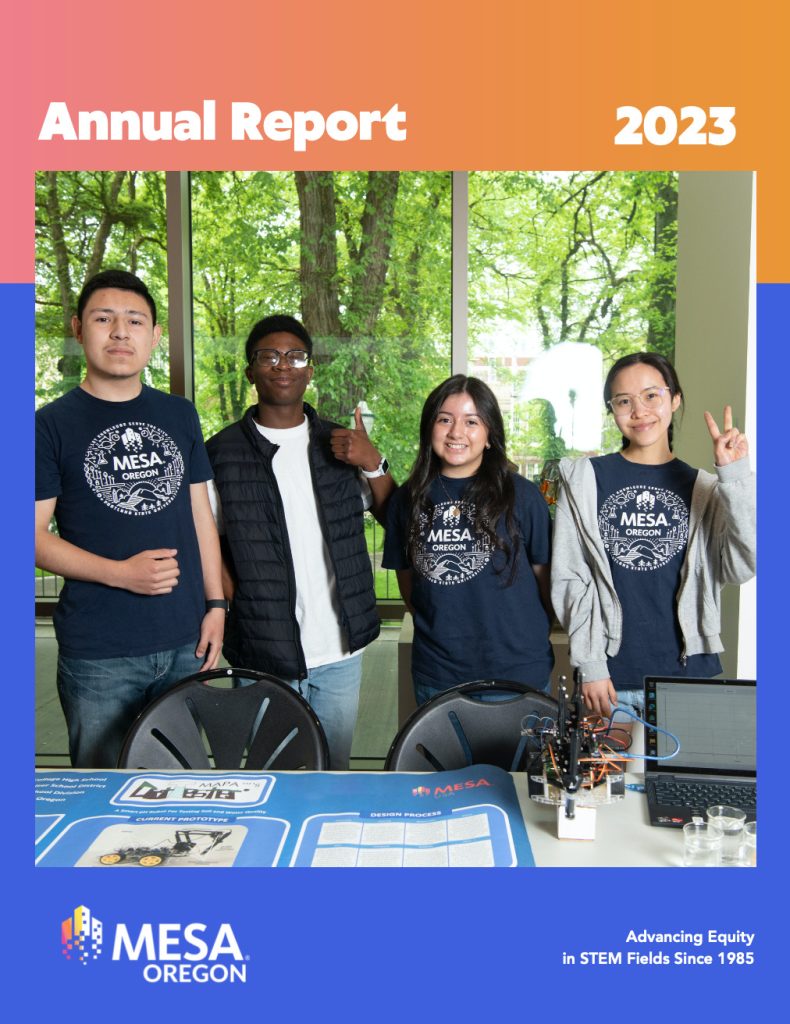 Four students stand behind a table displaying STEM projects and equipment, smiling in matching MESA Oregon shirts. The background shows large windows with green trees outside. Learn more in Our 2023 Annual Report.