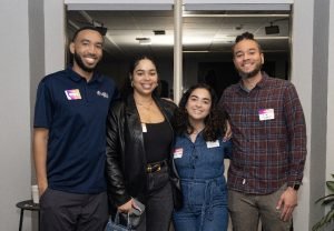 Four people stand close together indoors, smiling at the camera at our Celebrating Black Futures: Highlights from our latest Career Conversations Event. Three wear name tags, dressed casually—one in a leather jacket, another in a plaid shirt. A window is behind them.