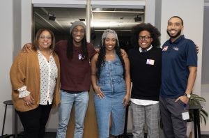 Five people stand indoors, smiling at the camera during our Celebrating Black Futures: Highlights from our latest Career Conversations Event. They are casually dressed, standing close together in a relaxed setting with a window and plants in the background.