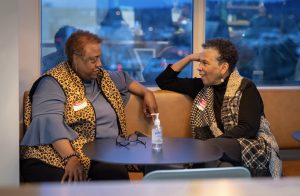 Two older women sit at a table by a window, engaged in conversation at our Celebrating Black Futures: Highlights from our latest Career Conversations Event. Both wear animal print vests, with hand sanitizer, glasses, and name tags on the table between them.