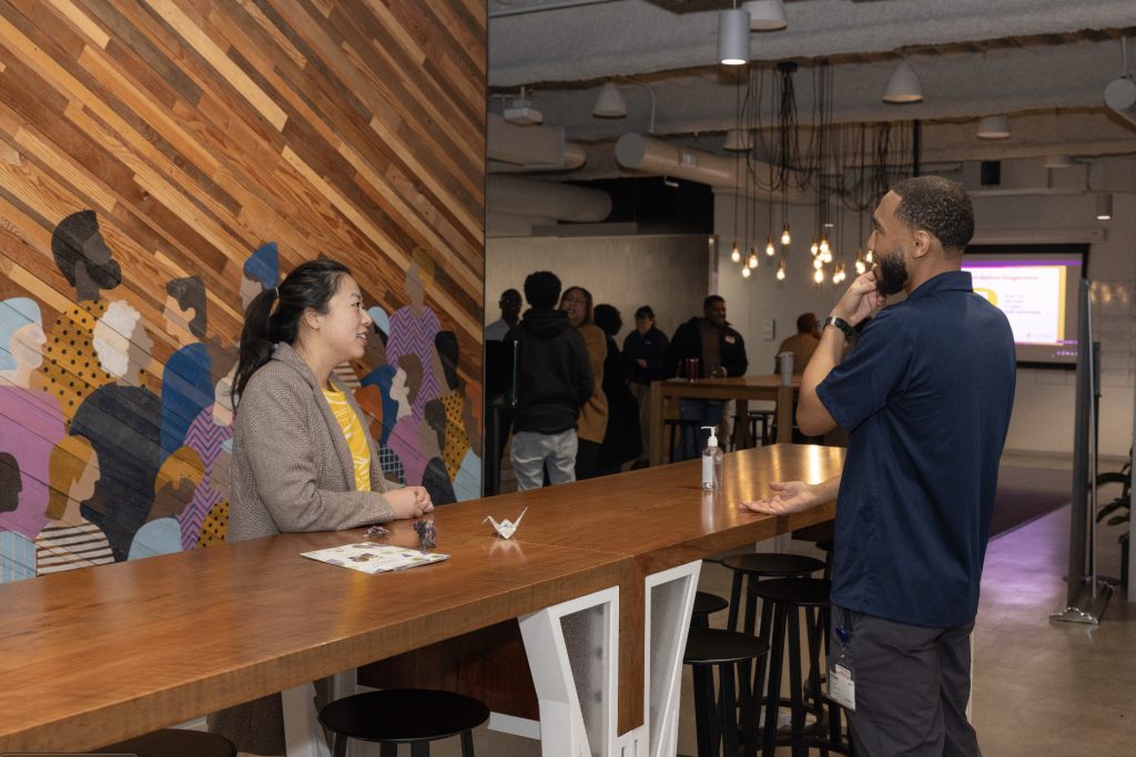 Two people have a conversation at a wooden high-top table in a modern office space, with a colorful mural of people on the wall behind them—captured as part of the demo day 2023 photo gallery.