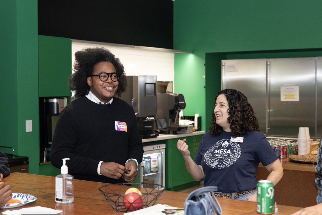 Two people stand and chat in a kitchen area in this demo day 2023 photo gallery. The man, wearing glasses and a black sweater, smiles while a woman in a blue Mesa Outdoors shirt gestures as she talks. A can of Sprite and hand sanitizer are on the counter.