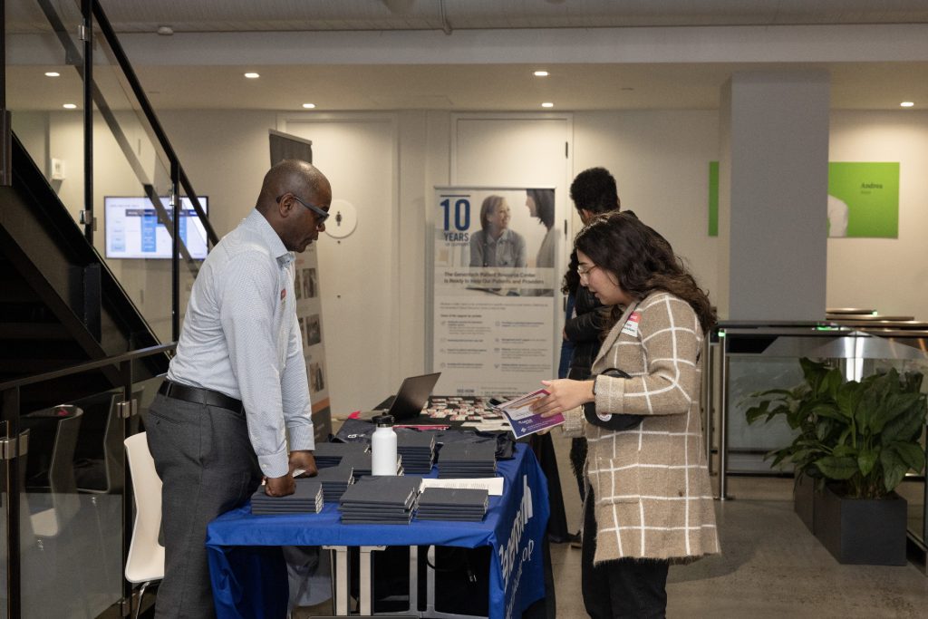 A man stands behind a table with pamphlets and booklets, speaking to a woman holding brochures at an indoor event or information fair—captured in this demo day 2023 photo gallery. Another person is visible in the background near informational posters.