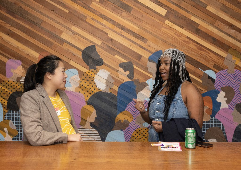 Two women sit at a wooden table, engaged in conversation. Behind them is a colorful mural of abstract people. This demo day 2023 photo gallery shot features one woman in a blazer, the other in a denim top with braided hair.
