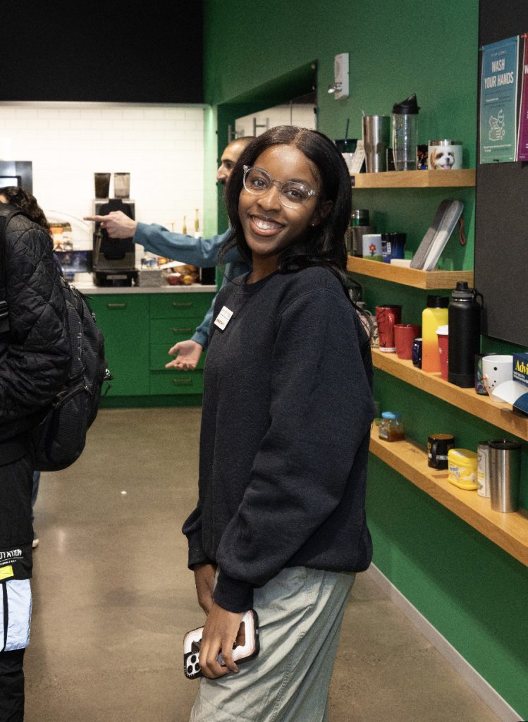 A woman with long dark hair, glasses, and a dark sweater smiles while standing indoors, holding a phone. Behind her are shelves with colorful mugs and a green wall—captured in this demo day 2023 photo gallery.