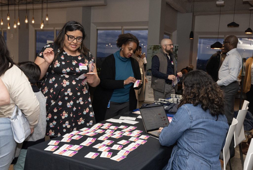 People gather at a registration table covered with name tags and a laptop during the demo day 2023 event. Some pick up name tags while others converse in the background under modern hanging lights, capturing moments for the photo gallery.