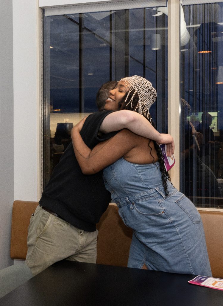 Two people warmly embrace by a window at night in this demo day 2023 photo gallery. One wears a sleeveless denim dress and a beaded headpiece, while the other wears a dark shirt and khaki pants. A brochure and phone rest on the black table nearby.