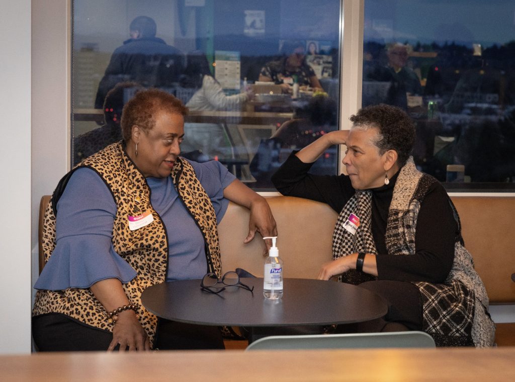 Two women sit at a round table by a window, deep in conversation. A bottle of hand sanitizer sits between them as they wear patterned vests and name tags. The evening cityscape shines outside—just one moment from our demo day 2023 photo gallery.