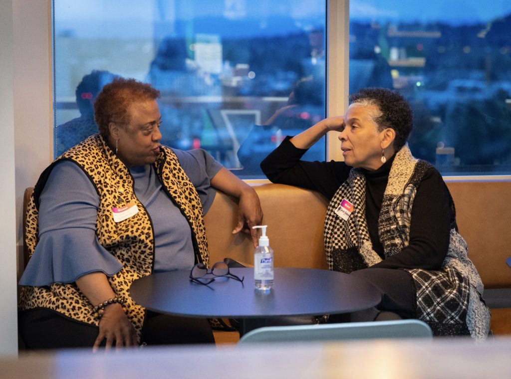 Two women sit at a round table near a window, engaged in conversation for the demo day 2023 photo gallery. Both wear patterned vests, with a bottle of hand sanitizer on the table. City lights shine outside.