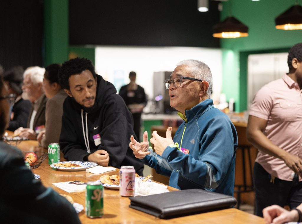 Two men sit at a wooden table engaged in conversation at Demo Day 2023, with snacks and drinks in front of them. Other people are seated and standing in the background of this brightly lit, modern photo gallery space.