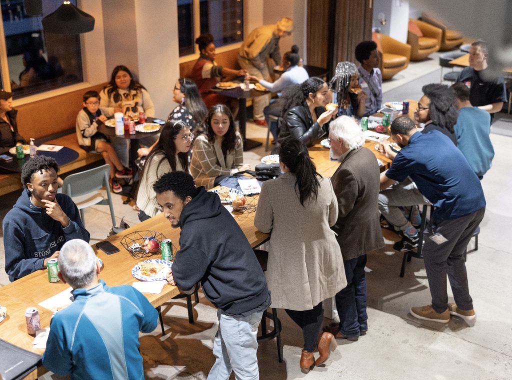 A diverse group of people sit and stand around two long tables, sharing food and drinks in a lively, indoor social gathering. This demo day 2023 photo gallery captures a friendly, relaxed atmosphere as guests eat, talk, and connect.