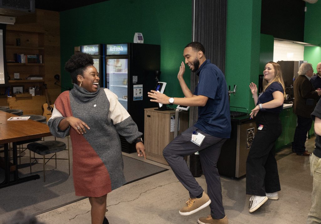 Three people are dancing and laughing together in a casual indoor setting with green walls and wooden tables. This lively, joyful moment is part of the demo day 2023 photo gallery, with vending machines and other guests in the background.